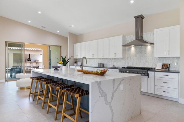 kitchen featuring vaulted ceiling, white cabinets, an island with sink, and wall chimney range hood