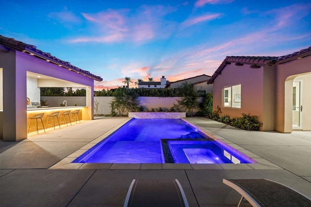 pool at dusk featuring an in ground hot tub, exterior bar, and a patio