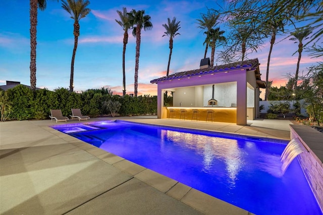 pool at dusk with pool water feature, a patio, and exterior bar