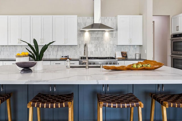 kitchen with a kitchen breakfast bar, wall chimney range hood, stainless steel double oven, light stone counters, and white cabinetry