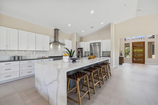 kitchen featuring wall chimney exhaust hood, dark stone countertops, appliances with stainless steel finishes, a large island, and a kitchen bar