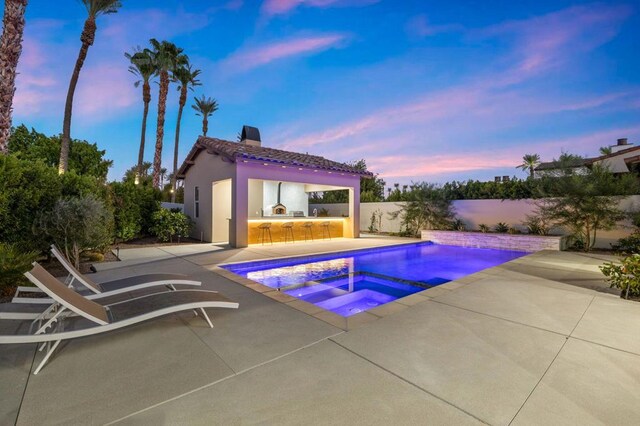 pool at dusk featuring an in ground hot tub, a patio area, and exterior kitchen