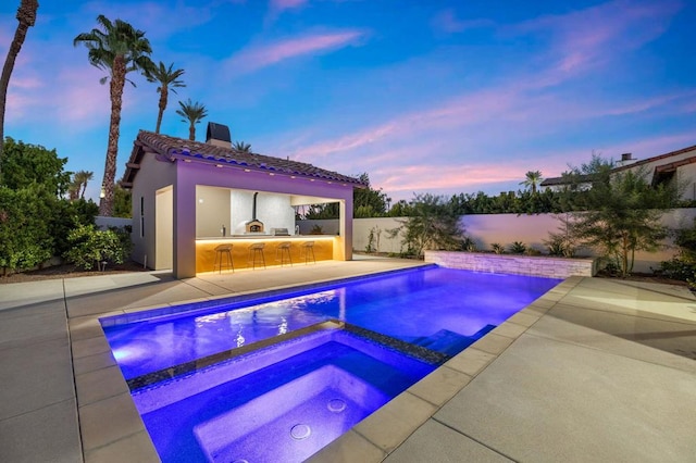 pool at dusk with an outdoor kitchen, a patio area, an outdoor bar, and an in ground hot tub