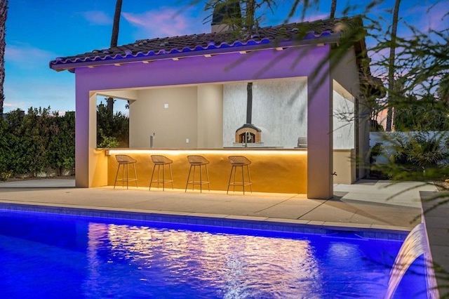 pool at dusk with pool water feature, a patio, and an outdoor bar