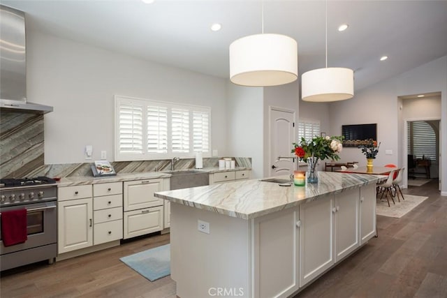 kitchen with light stone counters, wall chimney range hood, a center island with sink, stainless steel stove, and hanging light fixtures