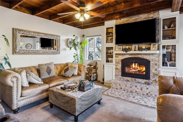 carpeted living room featuring built in features, beam ceiling, ceiling fan, a stone fireplace, and wooden ceiling