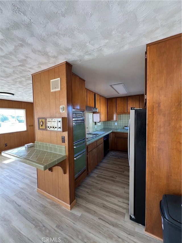 kitchen featuring tile countertops, visible vents, brown cabinetry, light wood-style floors, and black appliances