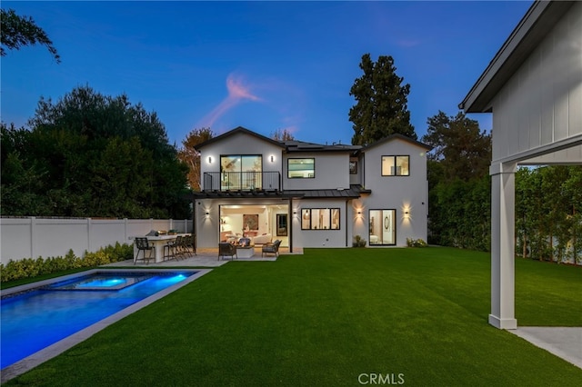 back house at dusk featuring an outdoor hangout area, a balcony, a pool with hot tub, a patio, and a lawn