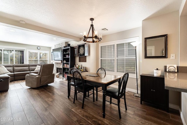 dining room with dark wood-type flooring, a stone fireplace, an inviting chandelier, a textured ceiling, and beamed ceiling