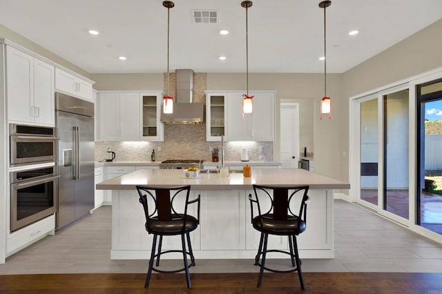 kitchen with stainless steel appliances, wall chimney exhaust hood, white cabinets, and an island with sink