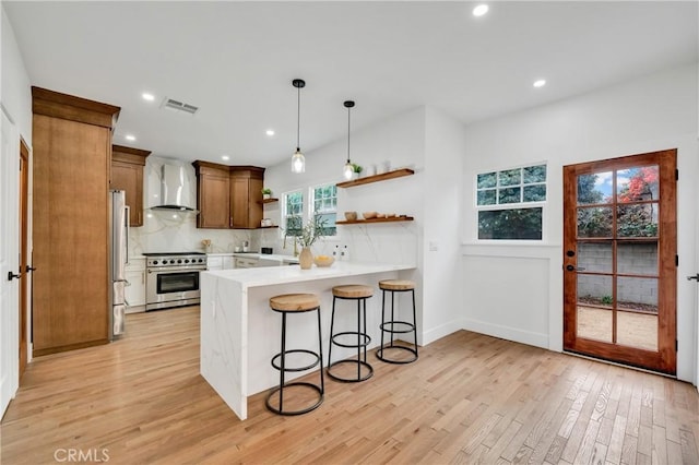 kitchen featuring kitchen peninsula, light wood-type flooring, stainless steel appliances, wall chimney range hood, and pendant lighting