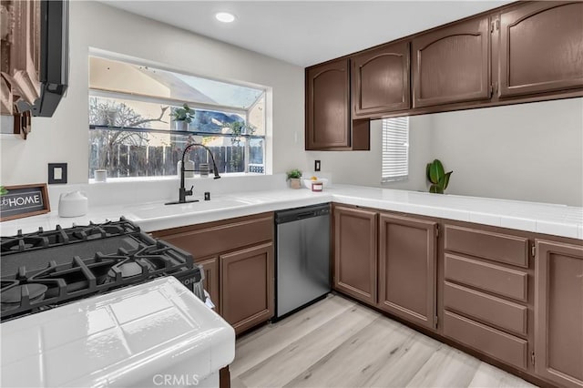 kitchen with sink, stainless steel dishwasher, tile counters, dark brown cabinets, and light wood-type flooring