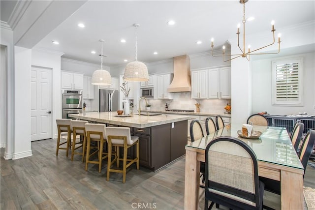 kitchen featuring stainless steel appliances, hanging light fixtures, a kitchen island with sink, white cabinets, and custom range hood