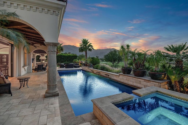 pool at dusk with a mountain view, ceiling fan, a patio area, and an in ground hot tub