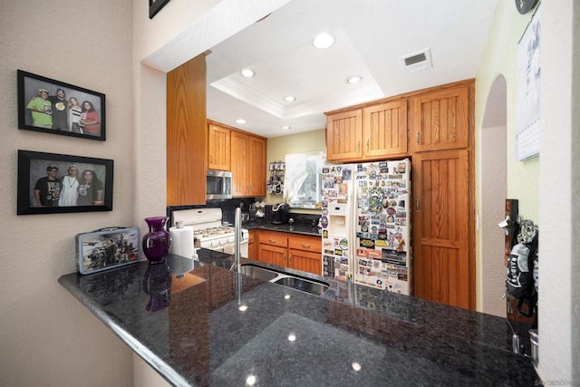 kitchen with white appliances, dark stone counters, sink, a raised ceiling, and kitchen peninsula