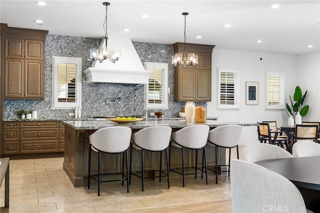 kitchen featuring hanging light fixtures, premium range hood, a center island with sink, and light stone counters