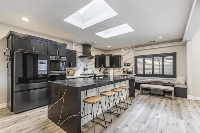 kitchen with a breakfast bar, a skylight, appliances with stainless steel finishes, an island with sink, and wall chimney exhaust hood