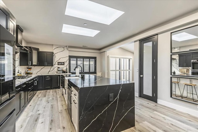 kitchen with tasteful backsplash, lofted ceiling with skylight, a center island with sink, light hardwood / wood-style flooring, and dark stone counters