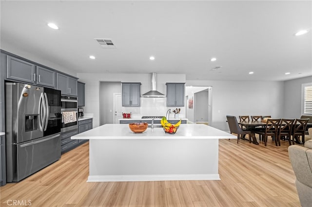 kitchen with stainless steel appliances, a center island with sink, wall chimney exhaust hood, and light hardwood / wood-style floors