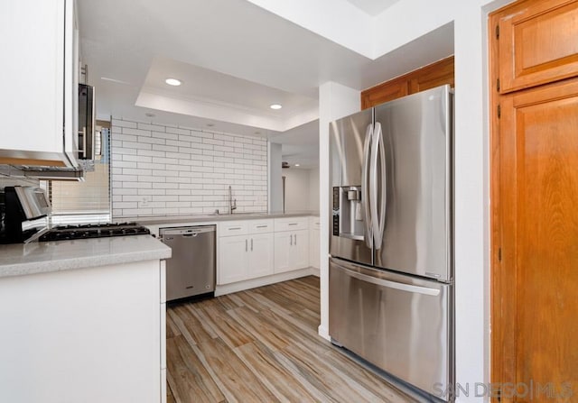 kitchen with white cabinets, a raised ceiling, light wood-type flooring, appliances with stainless steel finishes, and tasteful backsplash