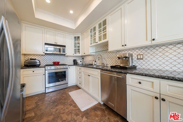 kitchen with appliances with stainless steel finishes, backsplash, dark stone counters, a raised ceiling, and sink