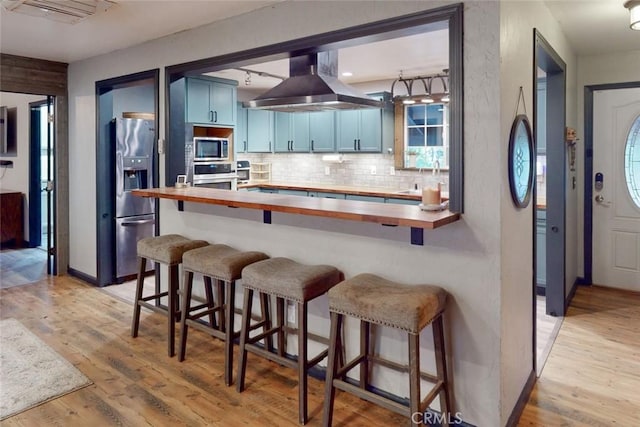 kitchen featuring a breakfast bar, ventilation hood, blue cabinetry, butcher block countertops, and stainless steel appliances