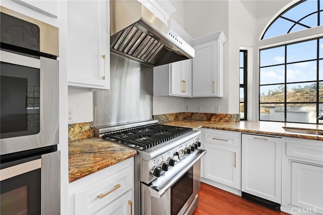 kitchen with stainless steel appliances, wall chimney range hood, dark stone countertops, white cabinets, and hardwood / wood-style floors