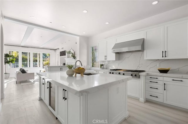 kitchen with backsplash, wall chimney range hood, sink, a center island with sink, and white cabinetry