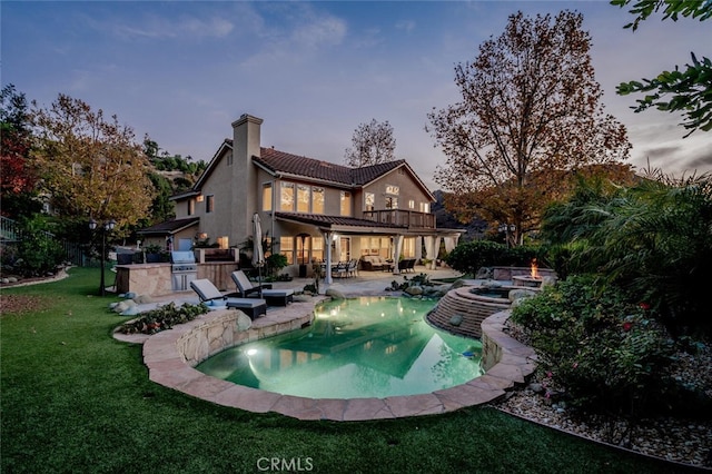 back house at dusk with an outdoor kitchen, a pool with hot tub, a balcony, a yard, and a patio area