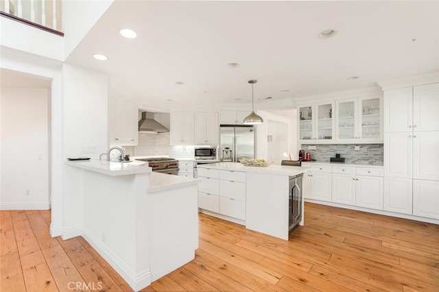 kitchen with appliances with stainless steel finishes, wall chimney range hood, light hardwood / wood-style flooring, white cabinetry, and hanging light fixtures