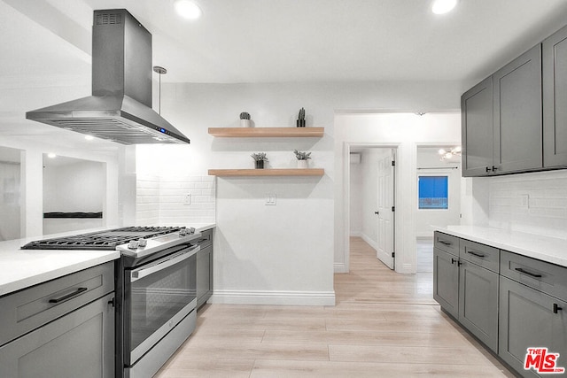 kitchen with gray cabinetry, backsplash, wall chimney exhaust hood, stainless steel gas stove, and light wood-type flooring