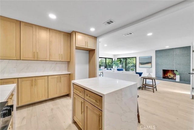 kitchen featuring light brown cabinetry, tasteful backsplash, a tile fireplace, a center island, and light hardwood / wood-style floors