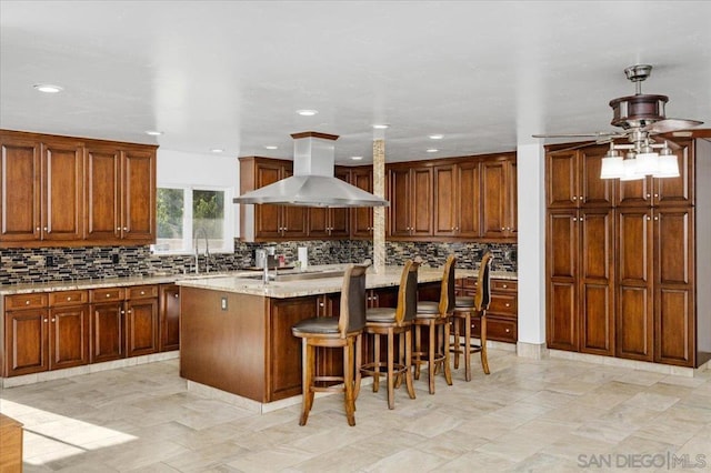 kitchen featuring a center island with sink, decorative backsplash, island range hood, and light stone counters