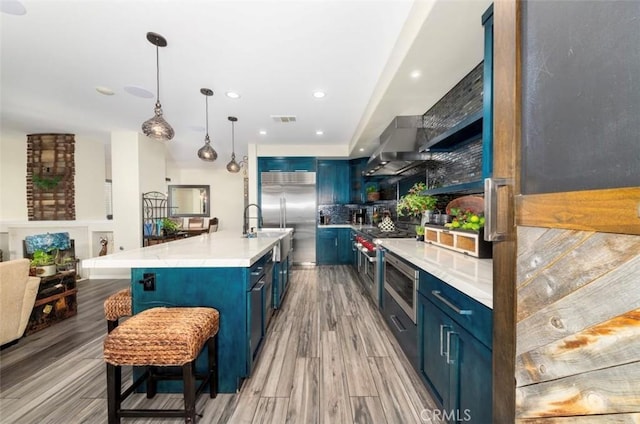 kitchen featuring high quality appliances, a center island with sink, blue cabinets, wall chimney exhaust hood, and light hardwood / wood-style floors