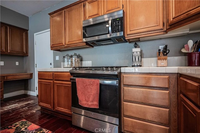 kitchen with brown cabinetry, dark wood-style flooring, tile counters, built in desk, and appliances with stainless steel finishes