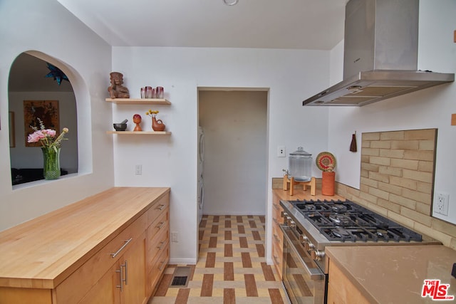 kitchen with high end range, wood counters, light brown cabinets, wall chimney range hood, and stacked washer and dryer