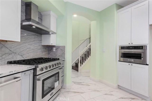 kitchen with backsplash, light stone counters, stainless steel appliances, wall chimney range hood, and white cabinetry