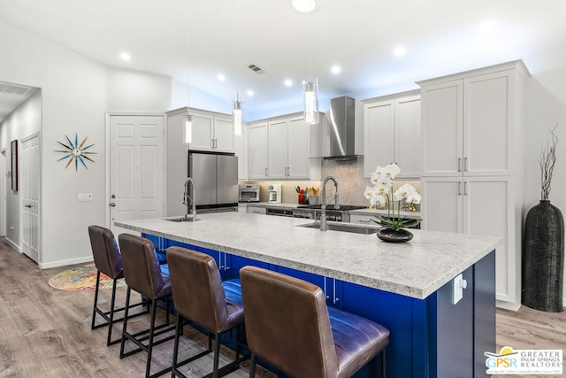 kitchen with lofted ceiling, a kitchen island with sink, hanging light fixtures, wall chimney exhaust hood, and stainless steel refrigerator