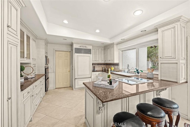 kitchen featuring a tray ceiling, a kitchen breakfast bar, dark stone counters, and a sink