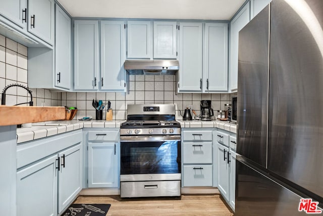 kitchen with tile countertops, stainless steel gas range, decorative backsplash, fridge, and light hardwood / wood-style floors