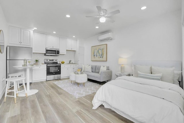 bedroom with an AC wall unit, light hardwood / wood-style floors, stainless steel refrigerator, ceiling fan, and sink