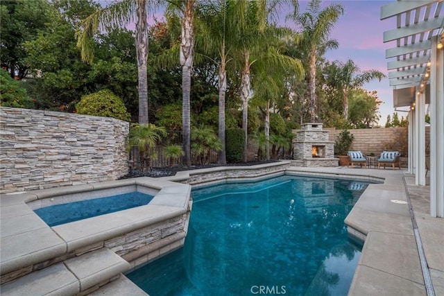 pool at dusk featuring an outdoor stone fireplace, a patio area, and an in ground hot tub