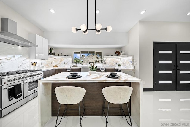 kitchen featuring wall chimney range hood, double oven range, hanging light fixtures, white cabinets, and a kitchen bar