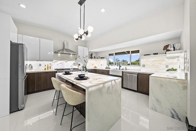 kitchen featuring wall chimney range hood, appliances with stainless steel finishes, white cabinetry, dark brown cabinets, and a kitchen island