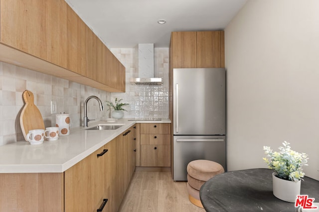 kitchen featuring stainless steel refrigerator, sink, wall chimney exhaust hood, decorative backsplash, and light wood-type flooring