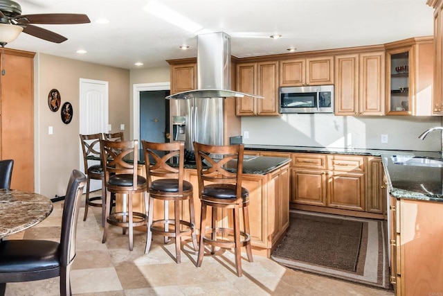 kitchen featuring a breakfast bar, sink, ceiling fan, dark stone countertops, and island exhaust hood