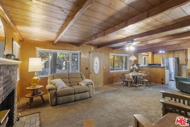 living room featuring wood walls, a brick fireplace, ceiling fan, beam ceiling, and carpet