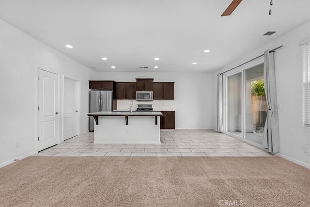 kitchen featuring appliances with stainless steel finishes, light colored carpet, ceiling fan, a breakfast bar area, and an island with sink