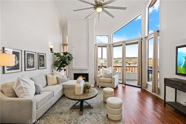 living room with ceiling fan, dark wood-type flooring, a high ceiling, a brick fireplace, and ornamental molding