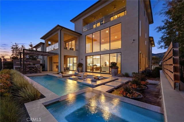 back house at dusk featuring a swimming pool with hot tub, a balcony, a patio area, and exterior kitchen